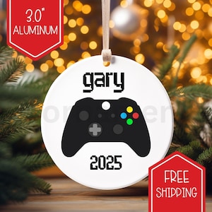 May include: A white, round Christmas ornament featuring a black video game controller. The name "gary" and the year "2025" are printed on the ornament. The ornament is 7.6 cm in diameter and made of aluminum.