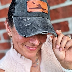 May include: A distressed charcoal gray baseball cap with a black mesh back and a rectangular tan leather patch on the front. The patch features a dark brown laser-engraved silhouette of a person paddling a kayak with an oar.