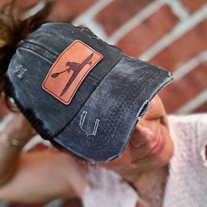 May include: Distressed black baseball cap with a brown leather patch featuring a kayak and kayaker design. The cap has a pre-curved bill and a ponytail opening. The hat has a worn, vintage look.