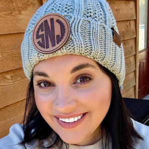 May include: A light gray knit beanie with a brown leather patch featuring the initials "SND". The beanie has a ribbed design and a small brown leather tag. A cozy winter accessory.