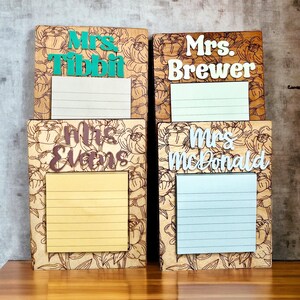May include: Four rectangular wooden memo boards with laser-engraved floral patterns. Each board features a personalized name in raised lettering and a lined notepad. The names are Mrs. Tibbit, Mrs. Brewer, Mrs. Evans, and Mrs. McDonald.