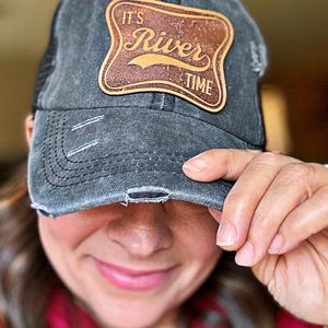 May include: Distressed gray baseball cap with a mesh back and a brown leather patch. The patch features the words "It's River Time" in a stylized font. The cap has a worn, vintage look with distressed details.