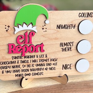 May include: A rectangular wooden sign titled Elf Report with a green and white elf hat graphic. It features the text Santa's making a list & checking it twice. I will report back every night, so he is gonna find out if you have been naughty or nice. Make good choices. It includes three white circular markers next to the labels Collins, Naughty, Almost There, and Nice.