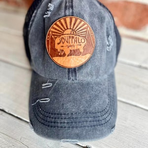 May include: A distressed, dark gray baseball cap with a black mesh back. The cap features a round, brown leather patch with the words "Buffalo River" and a graphic of a buffalo and sun.