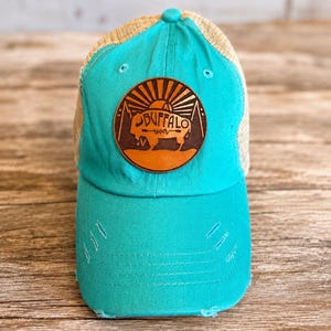 May include: A bright turquoise baseball cap with a distressed brim and a tan mesh back. A circular brown leather patch is centered on the front, featuring a laser-engraved graphic of a buffalo, mountains, a sunburst, and the text Buffalo River.