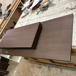 Pu&ograve; includere: Due tavole rettangolari in legno marrone scuro con una texture venata visibile. La tavola pi&ugrave; piccola &egrave; posizionata sopra quella pi&ugrave; grande, entrambe presentano bordi dritti e puliti e una superficie piatta e liscia color espresso.