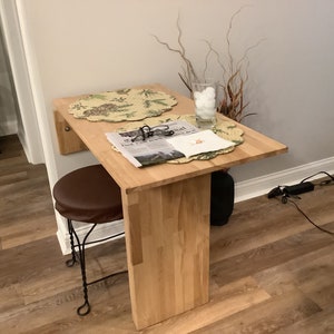 Pu&ograve; includere: Un tavolo pieghevole a parete in legno chiaro con piano rettangolare e una singola gamba di supporto verticale. Uno sgabello rotondo marrone con base in metallo &egrave; posizionato sotto. Due tovagliette circolari, un bicchiere con oggetti bianchi e un giornale sono appoggiati sopra.
