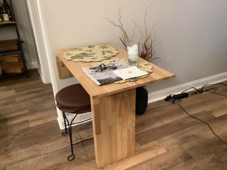 Pu&ograve; includere: Un tavolo pieghevole a parete in legno chiaro con piano rettangolare e una singola gamba di supporto verticale. Uno sgabello rotondo marrone con base in metallo &egrave; posizionato sotto. Due tovagliette circolari, un bicchiere con oggetti bianchi e un giornale sono appoggiati sopra.