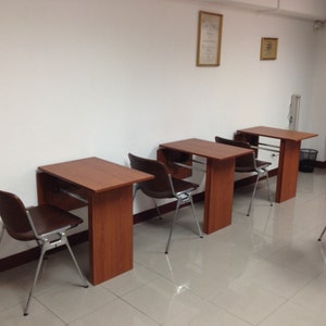 Pu&ograve; includere: Tre scrivanie individuali in legno con finitura marrone rossiccio, ciascuna abbinata a una sedia in plastica stampata marrone scuro con gambe in metallo argentato. Le scrivanie sono disposte in fila lungo una parete bianca.
