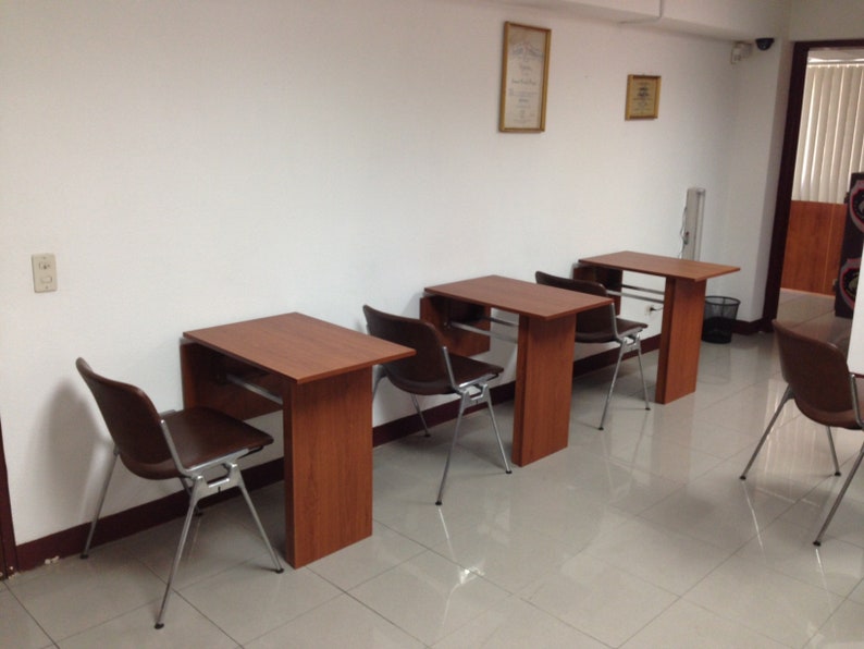 Pu&ograve; includere: Tre scrivanie individuali in legno con finitura marrone rossiccio, ciascuna abbinata a una sedia in plastica stampata marrone scuro con gambe in metallo argentato. Le scrivanie sono disposte in fila lungo una parete bianca.