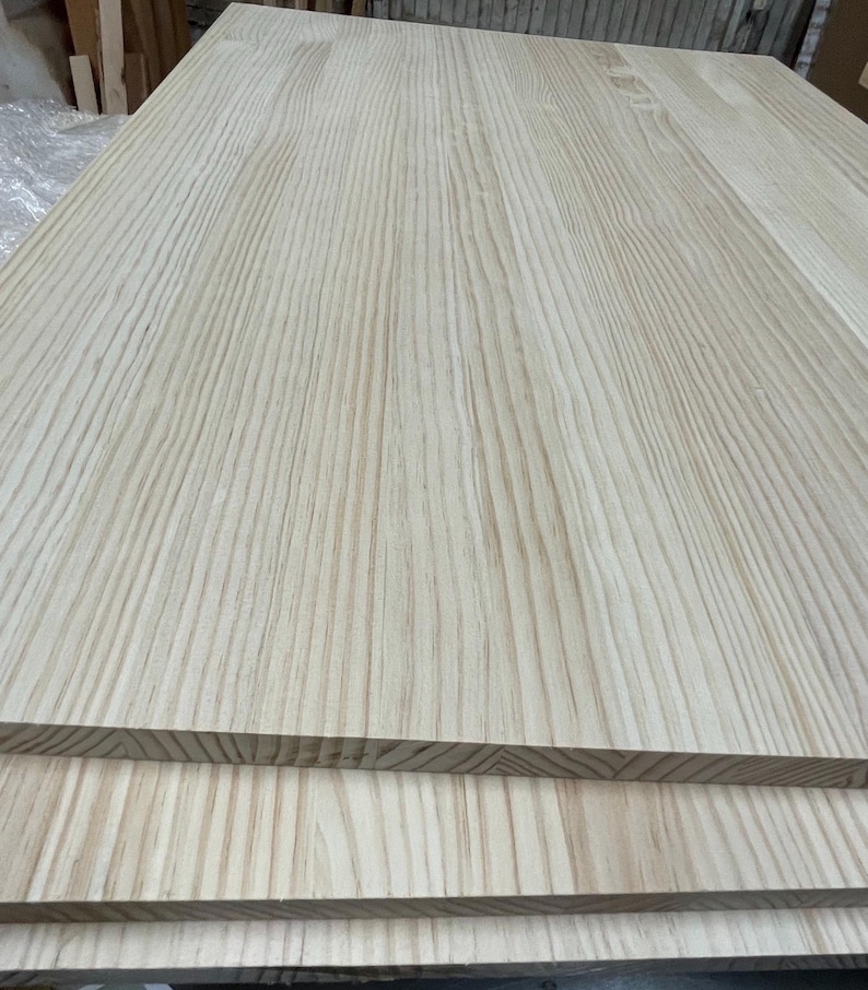 Pu&ograve; includere: Una pila di tavole di legno rettangolari di colore chiaro con un evidente motivo a venature verticali. Le tavole sono realizzate con pi&ugrave; strisce di legno unite, presentando una superficie liscia e non rifinita nei toni naturali del beige.