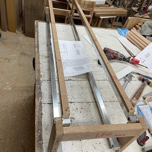 Puede incluir: Un marco de madera con barras de metal, etiquetado como "LEG BACK", se está ensamblando en un banco de trabajo. El marco está hecho de madera marrón claro y tiene un diseño simple.
