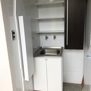 Op de afbeelding: Een witte wasbak met twee chromen kranen, een witte kast met twee deuren en witte planken boven de wasbak. De wasbak bevindt zich in een kleine, wit betegelde ruimte met een bruine kastdeur aan de rechterkant.
