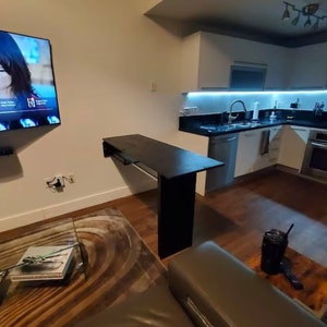 Pu&ograve; includere: Un tavolo rettangolare nero montato a parete con una singola gamba di supporto verticale, posizionato in uno spazio abitativo a pianta aperta tra una cucina con armadi bianchi e un soggiorno con televisione e divano grigio.