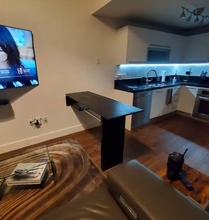 Pu&ograve; includere: Un tavolo rettangolare nero montato a parete con una singola gamba di supporto verticale, posizionato in uno spazio abitativo a pianta aperta tra una cucina con armadi bianchi e un soggiorno con televisione e divano grigio.