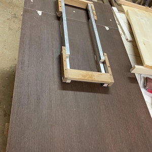 Pu&ograve; includere: Un pannello rettangolare in laminato con venature del legno marrone scuro, caratterizzato da un telaio centrale in metallo e legno chiaro. Il telaio &egrave; composto da due binari metallici argentati paralleli collegati da due traverse in legno chiaro.