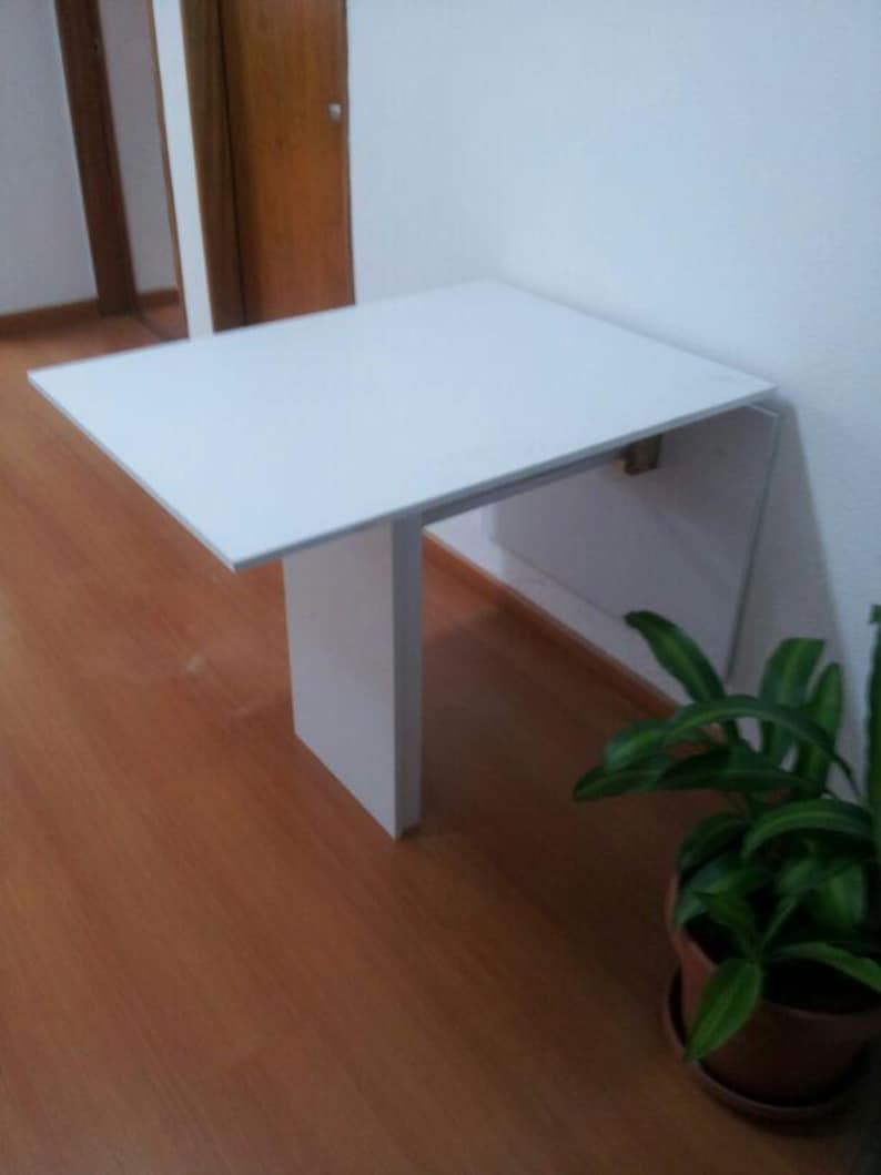 Pu&ograve; includere: Un tavolo pieghevole a parete di colore bianco con un piano rettangolare e una singola gamba di supporto verticale. Il tavolo &egrave; fissato a una parete bianca e si estende verso l'esterno, offrendo una superficie piana e liscia.