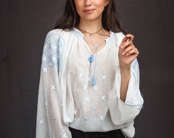 May include: A woman wearing a white embroidered blouse with blue accents and black jeans. The blouse has a V-neck and long sleeves with tassels.