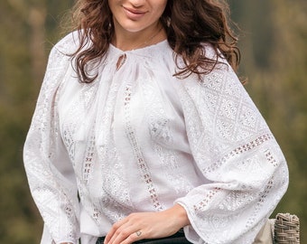 May include: A woman wearing a white embroidered blouse with long sleeves and black jeans sits on a wicker chair. The background is a blurred image of a forest.