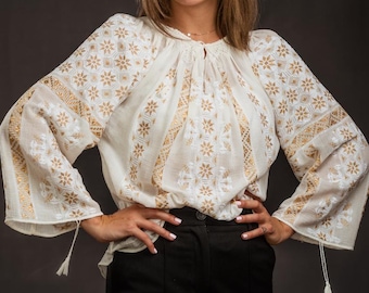 May include: A cream-colored long-sleeve blouse featuring intricate white and gold floral embroidery patterns, a gathered round neckline with a small front slit, and wide sleeves finished with delicate hanging tassels at the cuffs.