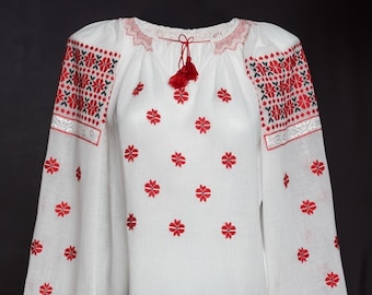 May include: White embroidered blouse with red floral patterns and black and white geometric designs on the sleeves. The blouse has a gathered neckline and a tassel tie.