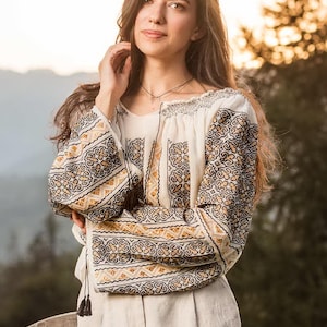 May include: Cream blouse featuring detailed black and gold embroidery on the sleeves and neckline. The traditional design is paired with light-coloured linen trousers. The overall look is elegant and classic, suitable for various occasions.