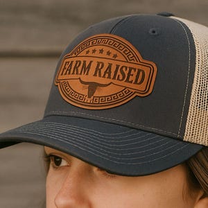 May include: A grey and beige trucker cap featuring a leather patch with the words "FARM RAISED" and a bull illustration. The cap has a curved peak and a mesh back. The image also includes the text "INSTANT DIGITAL DOWNLOAD".