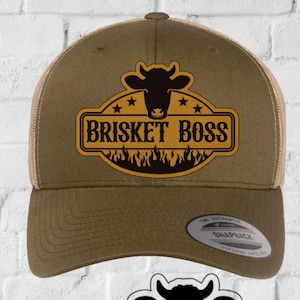 May include: A brown trucker cap with a mesh back and a curved peak. The cap displays a logo with a cow head, the words "BRISKET BOSS", and flames. The image also includes a digital download graphic.