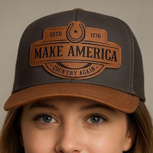 Pode incluir: Boné de caminhoneiro cinza e castanho com um patch de couro que diz "MAKE AMERICA COUNTRY AGAIN" com uma ferradura e o ano de 1776. O boné tem uma aba castanha e uma parte de trás em rede. A imagem também inclui o texto "INSTANT DIGITAL DOWNLOAD".