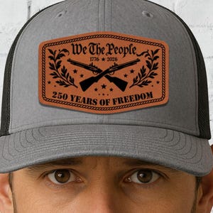 以下が含まれることがあります： 黒いメッシュの背面が付いたグレーのトラッカーハット。前面には、「We The People 1776-2026 250 YEARS OF FREEDOM」の文字と交差したライフルが書かれた茶色の革パッチが付いています。帽子は人がかぶっています。
