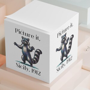 May include: A white cube-shaped notepad with a raccoon illustration and the text "Picture it, Sicily, 1912" on multiple sides. The raccoon is depicted in shades of gray and black, standing upright. The background features geometric shapes in pastel colors.