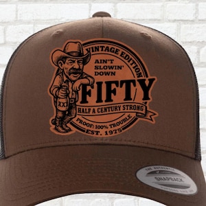 May include: Brown trucker hat with a mesh back and a snap closure. The front features a vintage-style graphic with the text "FIFTY" and a cowboy illustration. The hat also includes the text "Ain't Slowin' Down" and "Half a Century Strong."