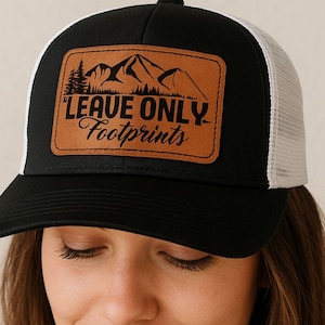 May include: Black trucker hat with a white mesh back and a curved brim. The front features a brown leather patch with the text "LEAVE ONLY Footprints" and a mountain and tree design. The hat is a digital download.