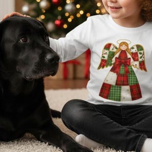 May include: A white long-sleeved shirt featuring a Christmas angel design with a red bow and patchwork-style dress. The angel has a gold halo and wings. The shirt is worn by a child sitting on a cream-colored rug next to a black dog.