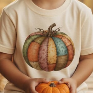 May include: A cream-colored t-shirt featuring a colorful, patchwork-style pumpkin design. The pumpkin has various shades of green, orange, pink, and blue. A small, orange pumpkin is held in the hands.