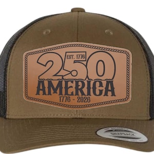 Puede incluir: Gorra trucker marrón con parte trasera de malla y cierre snapback. La parte delantera presenta un parche de cuero con el texto "250 AMERICA 1776-2026" y "EST. 1776". La gorra es de estilo clásico, adecuada para uso informal.