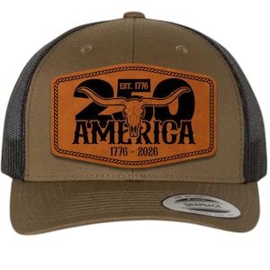 Puede incluir: Gorra de camionero marrón con parte trasera de malla y visera curva. La parte delantera presenta un parche de cuero con el texto "250 AMERICA 1776-2026" y un gráfico de calavera de cuernos largos. Cierre snapback.
