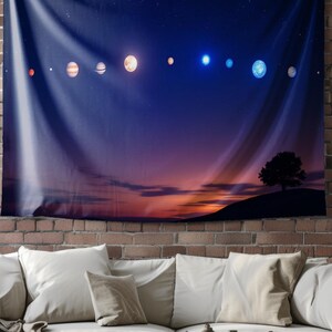 May include: A tapestry featuring a dark blue night sky with a line of planets and stars. The planets are a mix of orange, brown, blue, and white. The sky fades to a pink and purple sunset at the bottom of the tapestry. A silhouette of a tree is on a hill in the foreground.