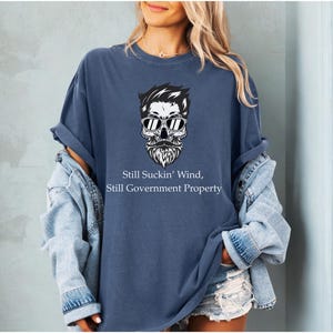 May include: A navy blue t-shirt with a skull graphic wearing sunglasses and a beard. The text on the shirt reads "Still Suckin' Wind, Still Government Property". The shirt is worn with a denim jacket and shorts.