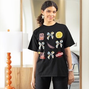 May include: A black t-shirt with a graphic design featuring a lemon, fish, bows, and cans of sardines. The text "BEST QUALITY" is printed on the shirt.