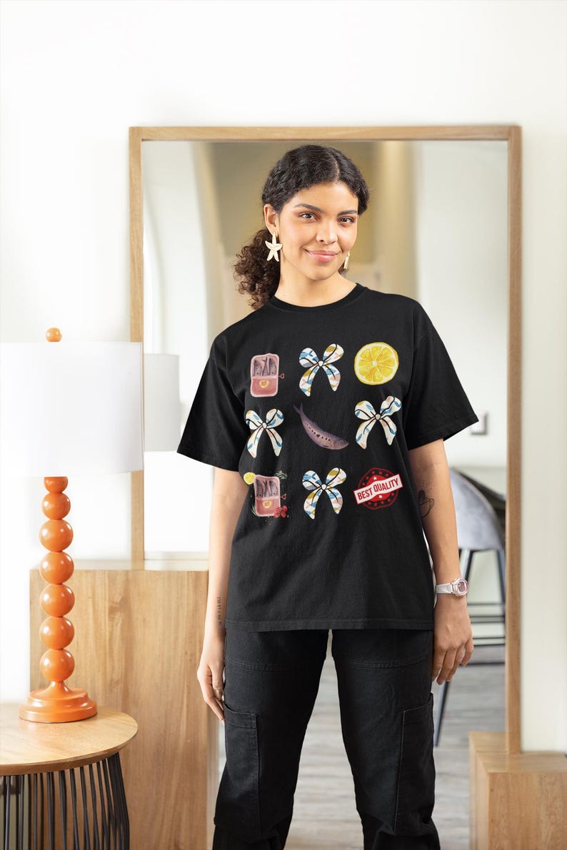 May include: A black t-shirt with a graphic design featuring a lemon, fish, bows, and cans of sardines. The text "BEST QUALITY" is printed on the shirt.
