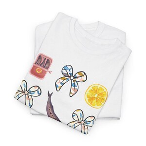 May include: White t-shirt featuring a colourful illustration of a fish, a lemon, and bows. The illustration is in a watercolour style. The t-shirt is folded in half.