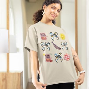 May include: A beige t-shirt featuring a variety of colourful illustrations, including a tin of sardines, lemons, bows, and the text "Best Quality".