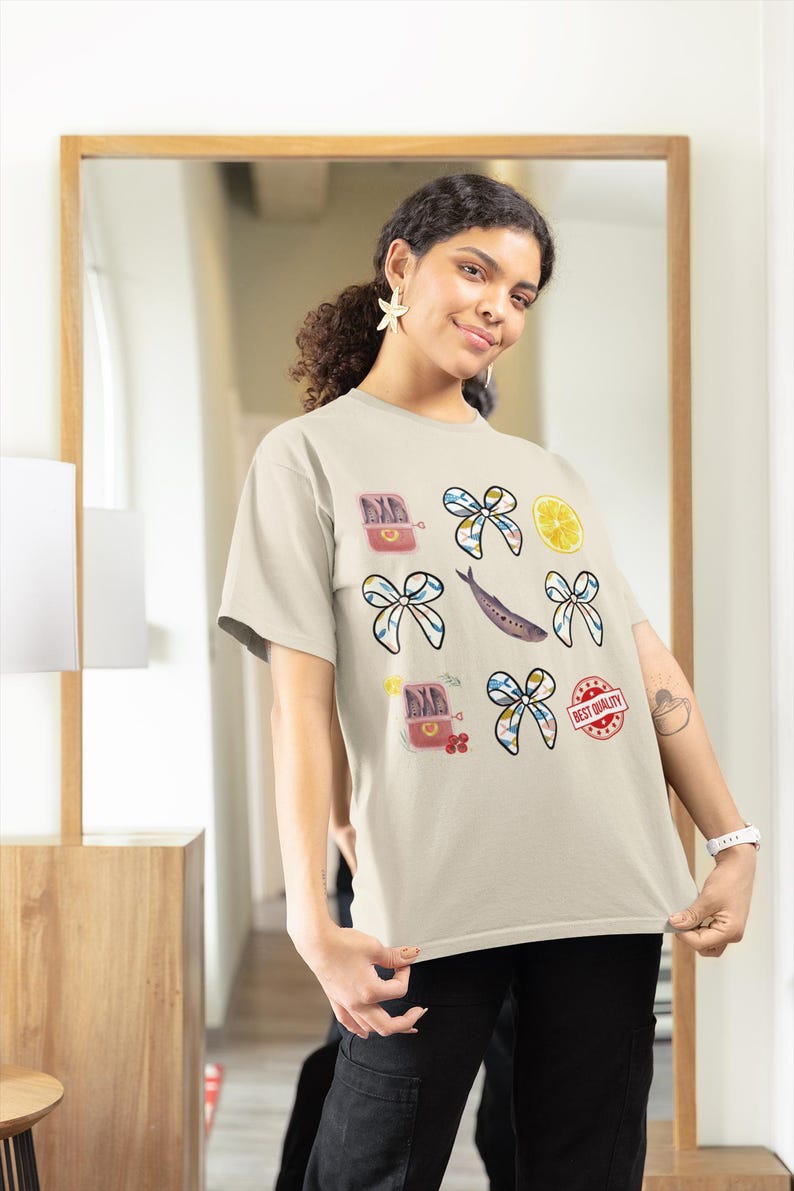 May include: A beige t-shirt featuring a variety of colourful illustrations, including a tin of sardines, lemons, bows, and the text "Best Quality".