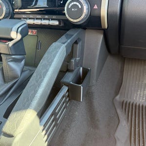 May include: Black plastic car interior accessory, likely a storage bin or organizer, mounted near the center console. The accessory is rectangular and appears to be designed to fit into the vehicle's existing structure. The image shows a close-up view of the item.