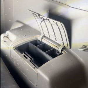 May include: A beige plastic automotive storage compartment with an open hinged lid, revealing a dark gray interior divided into three distinct sections for organizing small items within a vehicle interior console or dashboard area.