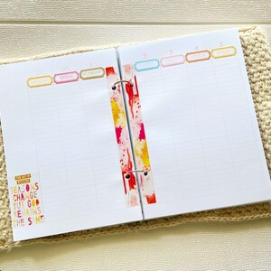 Puede incluir: Agenda abierto con páginas rayadas, etiquetas de días coloridas y un lomo decorativo. El planificador presenta el texto "The Art of Noticing" y la cita "Seasons Change But God Remains The Same."