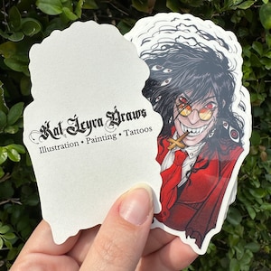 May include: A stack of white stickers with a black and white illustration of a vampire with red eyes and a cross. The text "Kat Leyva Draws" is written in black cursive on the sticker. The text "Illustration. Painting. Tattoos" is written in black below the name.
