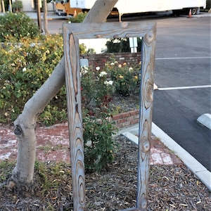 May include: A tall, rectangular mirror with a rustic wooden frame. The frame is painted a light blue color with a distressed finish. The mirror is reflecting a view of a garden with flowers and greenery.