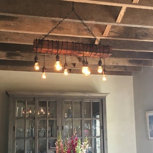 May include: A rustic wooden chandelier with exposed Edison bulbs hangs from a chain in a dining room with a wooden ceiling. The chandelier is made of a reclaimed wood beam with wire running through it. The chandelier is over a dining table with chairs and a hutch.